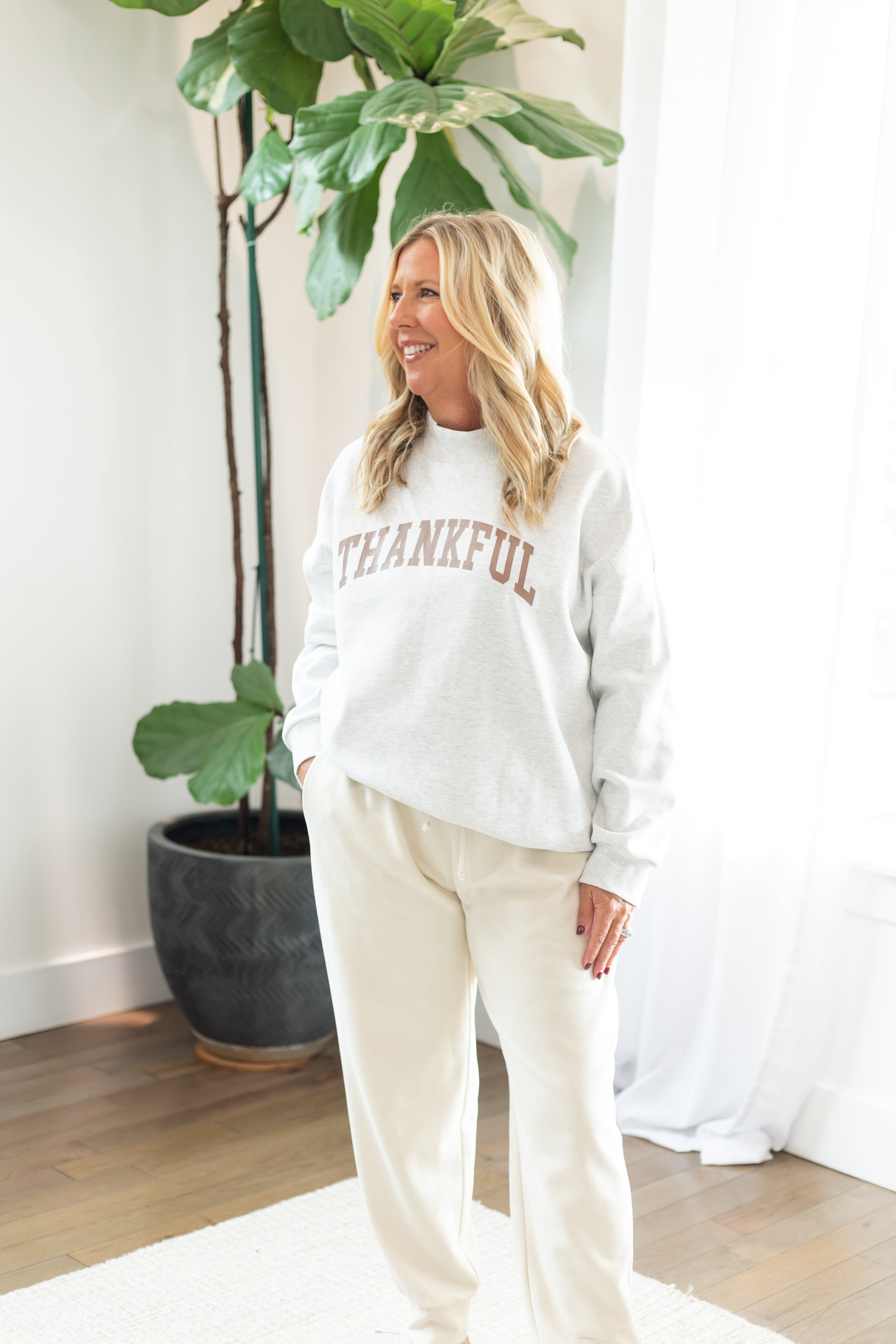 Woman wearing a white sweatshirt with 'THANKFUL' text, standing indoors with plants in the background.