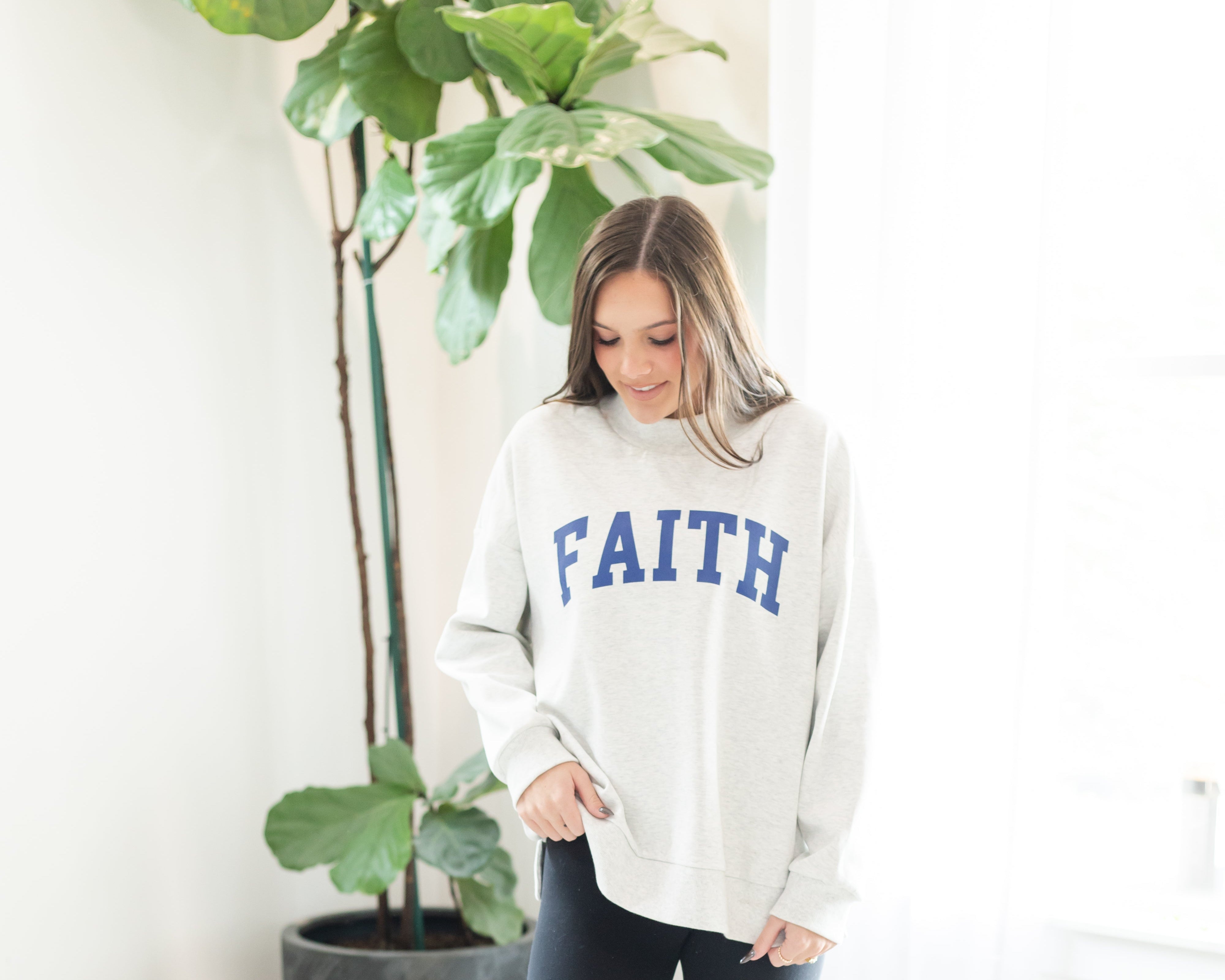 Woman wearing a white sweatshirt with 'FAITH' printed on it, standing in a room with a plant and white wall.