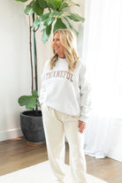Woman wearing a white sweatshirt with 'THANKFUL' text, standing indoors with plants in the background.