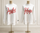 Two white crewneck sweatshirts displayed on mannequins against a light paneled wall. The left sweatshirt has the word 'Joyful' in red script across the chest, and the right sweatshirt has the word 'Hope' in red script across the chest.