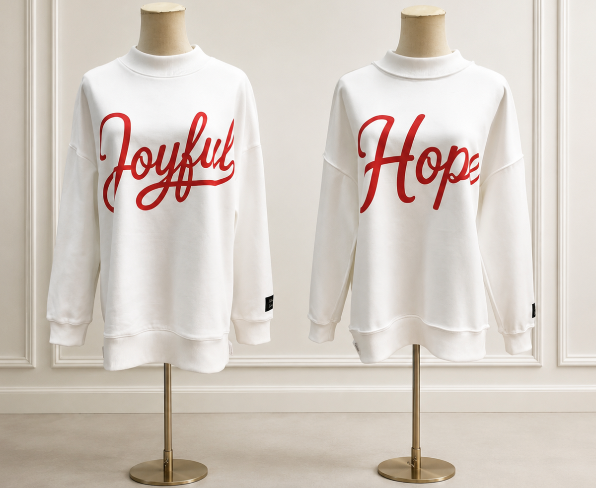 Two white crewneck sweatshirts displayed on mannequins against a light paneled wall. The left sweatshirt has the word 'Joyful' in red script across the chest, and the right sweatshirt has the word 'Hope' in red script across the chest.